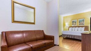 A hotel room with a brown leather sofa, a wooden table, a bed with white bedding, a wooden headboard, and two wall-mounted paintings. - 1 King Bed|Non-Smoking,Kitchen (Myrtle Beach SC)