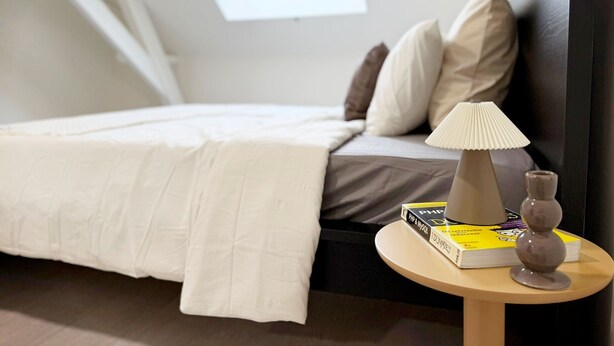 Una cama bien hecha con edredón blanco, una lámpara de noche, una mesita con libros y un florero, y una ventana con vistas.
