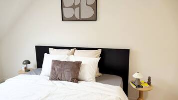 A neatly made bed with white bedding, a black headboard, and a few pillows. There is a framed abstract artwork above the bed, and a small round table with a lamp and a book on the side.