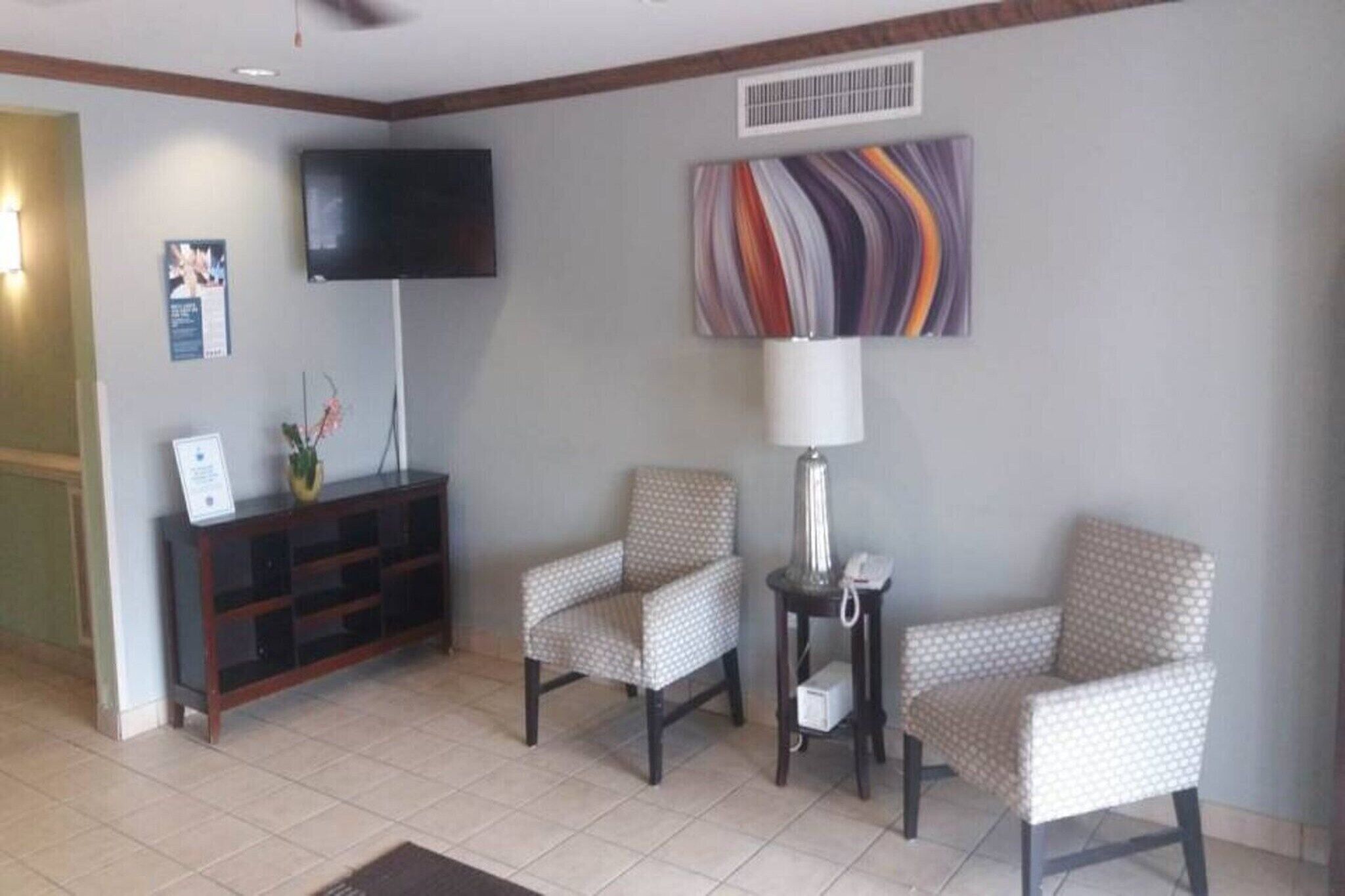 A hotel lobby with a television, a patterned wall art piece, a lamp, and two patterned chairs.