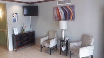 A hotel lobby with a television, a patterned wall art piece, a lamp, and two patterned chairs.