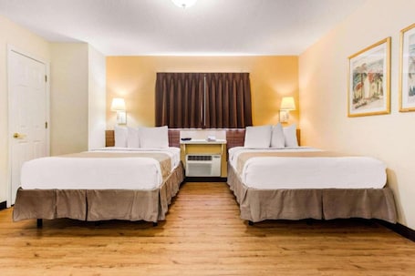 A hotel room with two beds, a desk, a chair, a lamp, and a wall-mounted air conditioner.