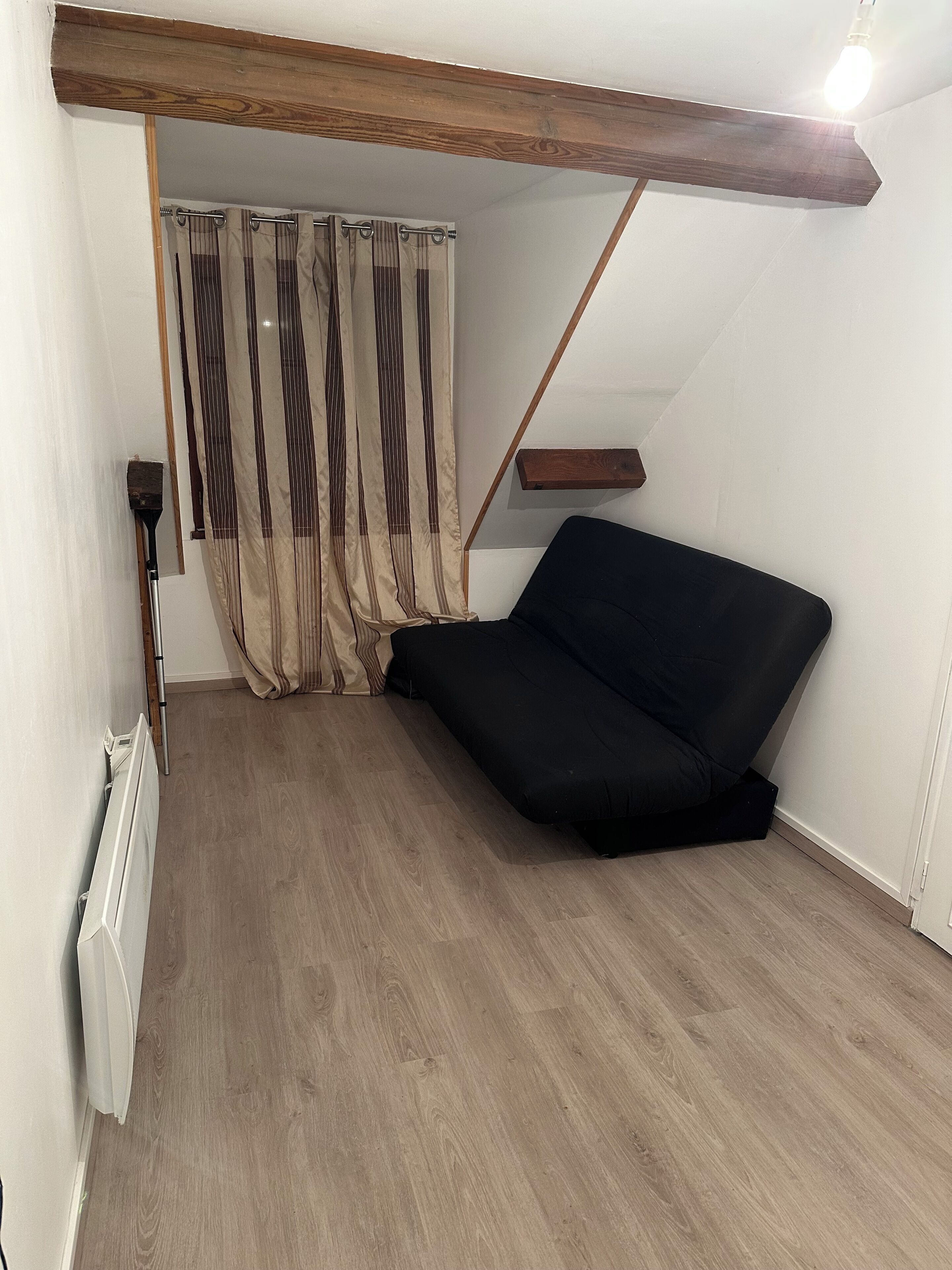 Una habitación con viga de techo de madera, un sofá negro, un radiador y una cortina.