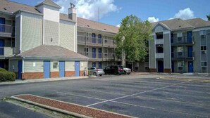 A multi-story motel with a parking lot, a small building with a sloped roof, and a tree in front.