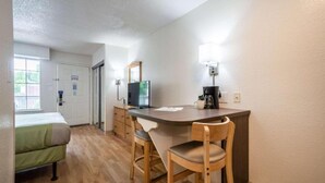 Une chambre d’hôtel de taille réduite, équipée d’un lit, d’un bureau avec un ordinateur, d’une chaise et d’une machine à café.