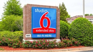 Une enseigne pour le Studio 6 Extended Stay, avec un symbole Wi-Fi, entourée de verdure et d’un mur de briques.