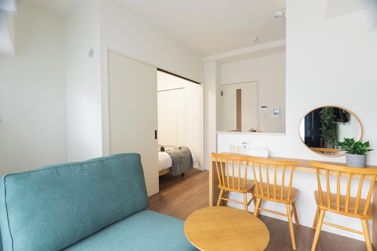 A modern room with a teal sofa, wooden chairs, a small round table, a bed, and a mirror on the wall.