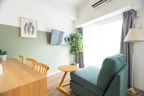 A room with a wooden dining table, chairs, a green sofa, a small round table, a potted plant, a TV on the wall, and a floor lamp. - Premiage Minoshima (Hakata Ward, Fukuoka)