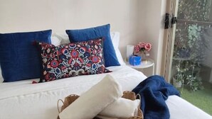 Una cama con ropa blanca, almohadas azules y una manta azul. Hay una cesta de mimbre con toallas dobladas y una mesita con un jarrón de flores.