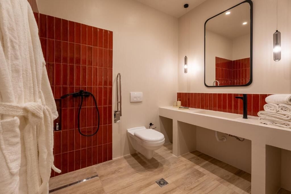 A modern bathroom with a red-tiled shower wall, a white toilet, a sink with a mirror, and a towel rack.