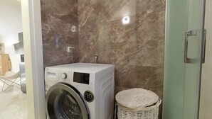 A modern laundry room with a front-loading washing machine, a marble-tiled wall, and a wicker basket. - Charming 3-Bedroom House Rental in Málaga, Spain (Málaga)