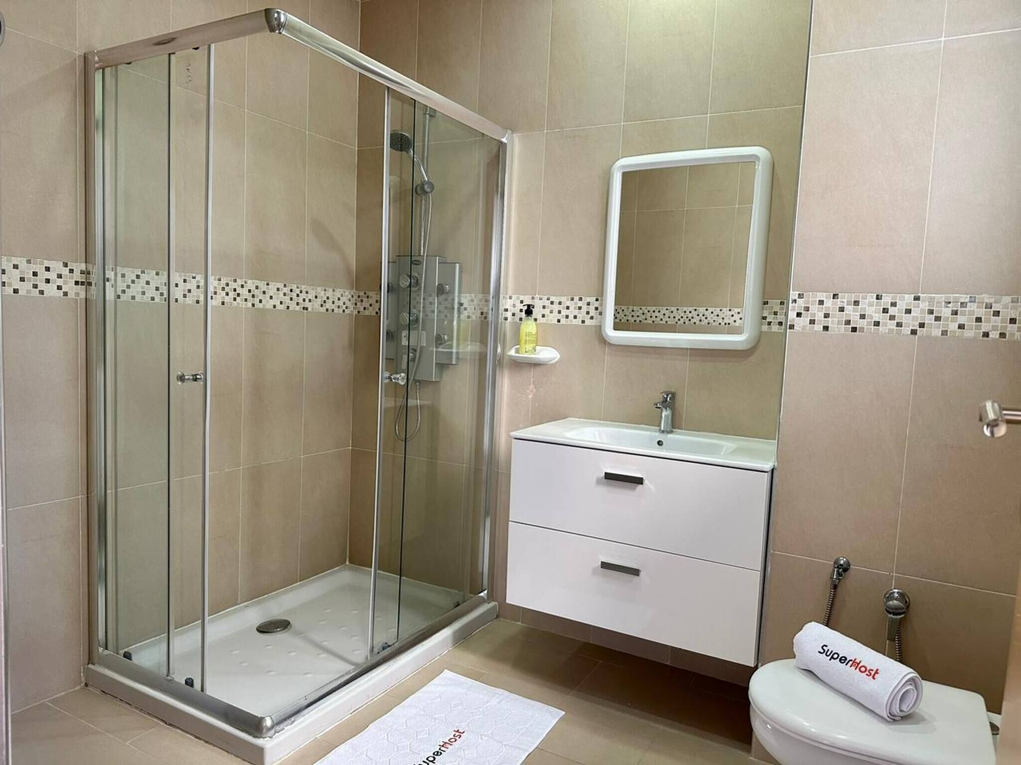 Ein modernes Badezimmer mit einer gläsernen Duschkabine, einem weißen Waschbecken mit Spiegel darüber und einer Toilette. Auf dem Handtuch ist „Superhost“ aufgedruckt.