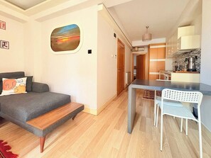 Ein modernes Wohnzimmer mit grauem Sofa, einer Holzbank, einem Esstisch mit weißem Stuhl und einer Küchenzeile mit Holzschränken und einem Wasserkocher.
