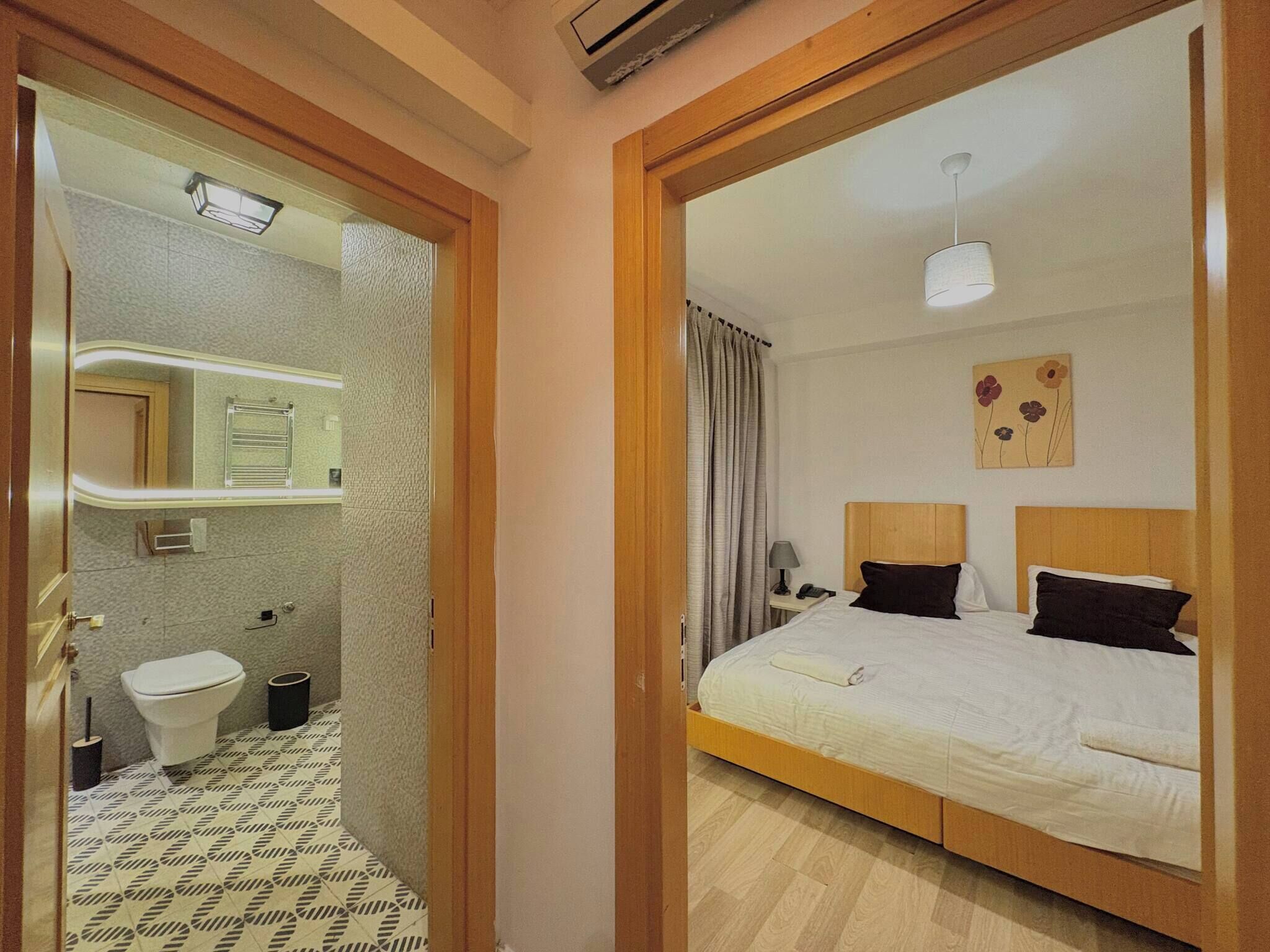Ein Hotelzimmer mit einem hölzernen Bett, einem Nachttisch mit Lampe, einem Badezimmer mit Toilette, einem Spiegel und einer Duschkabine.