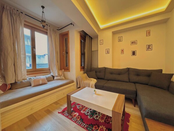 Een woonkamer met een bank, een hock met kussens, een salontafel en een kamerkleed met patroon.