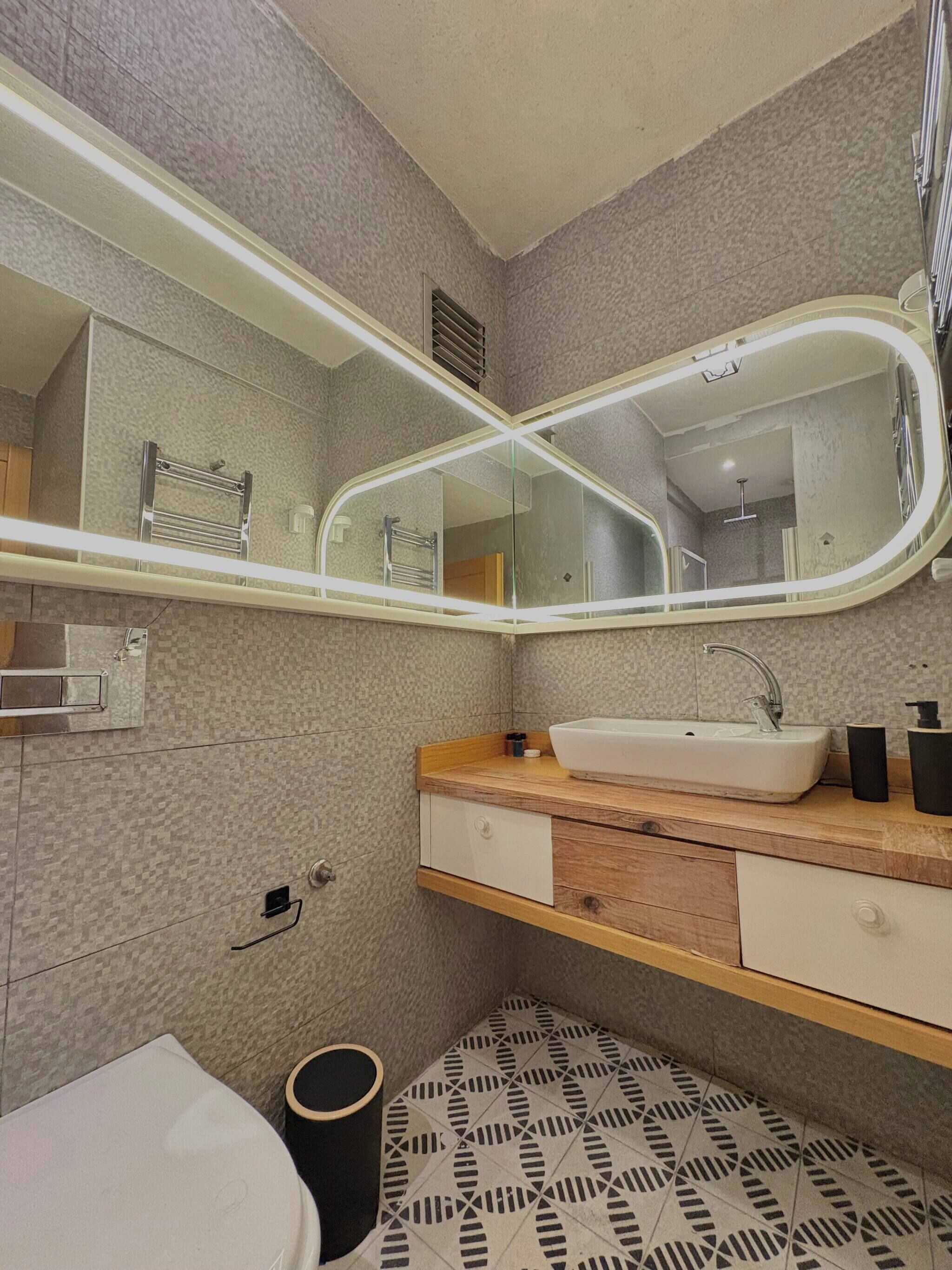 A bathroom with a white sink, wooden vanity, a large mirror with lights, patterned floor tiles, and a black trash bin.