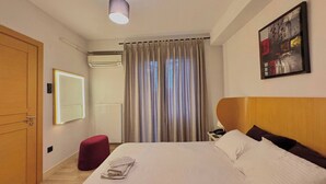 Une chambre d’hôtel avec un lit, un pouf rouge, un miroir fixé au mur, un appareil de climatisation, un luminaire au plafond et un tableau accroché au mur.