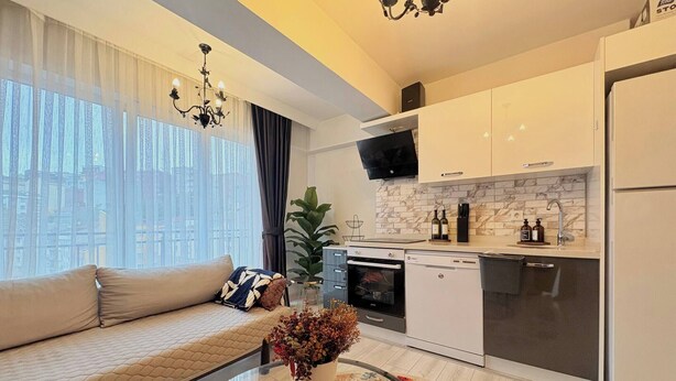 Un appartement moderne comprenant un canapé, une table basse en verre, une cuisine avec des meubles blancs, un four encastré et une grande fenêtre agrémentée de rideaux légers.