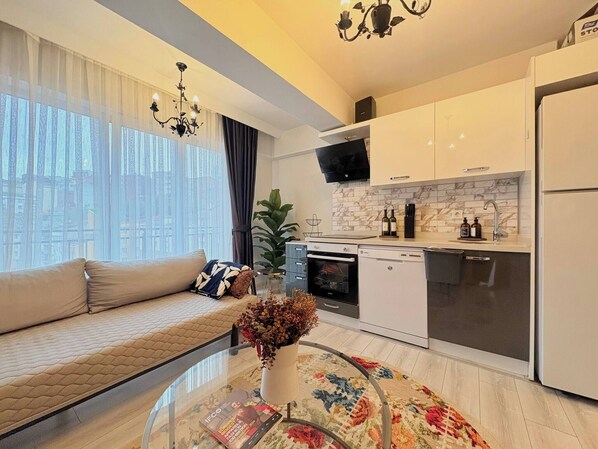 A modern apartment with a sofa, a glass coffee table, a kitchen with white cabinets, a built-in oven, and a large window with sheer curtains. - Bright & Modern 2BR Apartment in Kağıthane (Kağıthane)