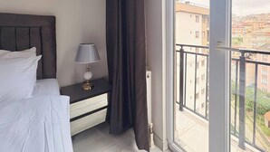 Une chambre d’hôtel avec un lit, une table de chevet avec une lampe, une fenêtre donnant sur des immeubles et un rideau.