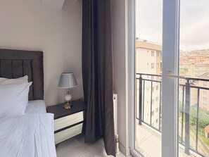 A hotel room with a bed, a nightstand with a lamp, a window with a view of buildings, and a curtain. - Bright & Modern 2BR Apartment in Kağıthane (Kağıthane)