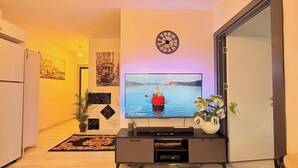 Un salon avec un téléviseur à écran plat, une horloge, une plante en pot et un tapis décoratif.