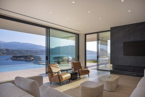 Ein modernes Wohnzimmer mit einem großen Glasfenster, das auf einen Balkon mit Blick auf Berge und Wasser führt.