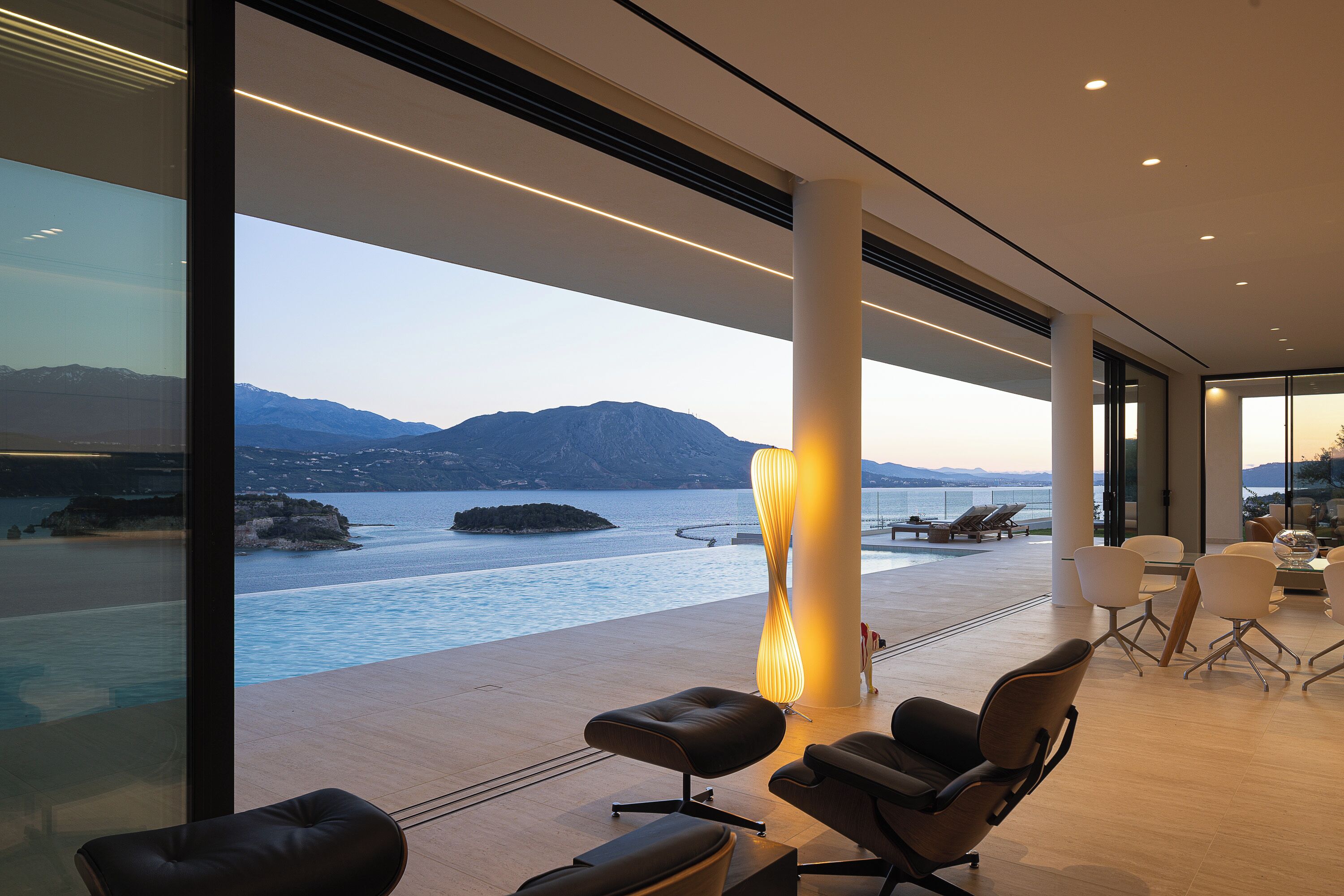 A modern interior with a view of a body of water and mountains, featuring a seating area with chairs and a table.