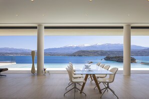 Ein moderner Essbereich mit Glastisch und Stühlen, große Fenster mit Blick auf eine Küstenlandschaft mit Bergen und einem Gewässer.