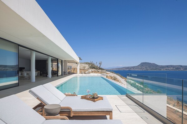 Ein moderner Poolbereich im Freien mit Liegestühlen, einem Holztisch mit Schale und Früchten sowie Blick auf das Meer und die Landschaft.