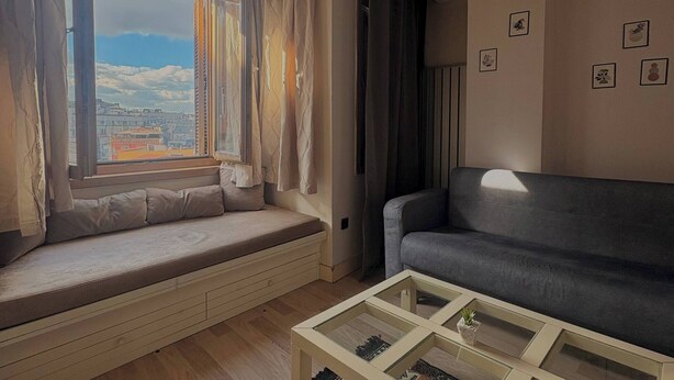 Ein Wohnzimmer mit einer Couch, einem Glastisch, einem Fenster mit Vorhängen und Blick auf Gebäude.
