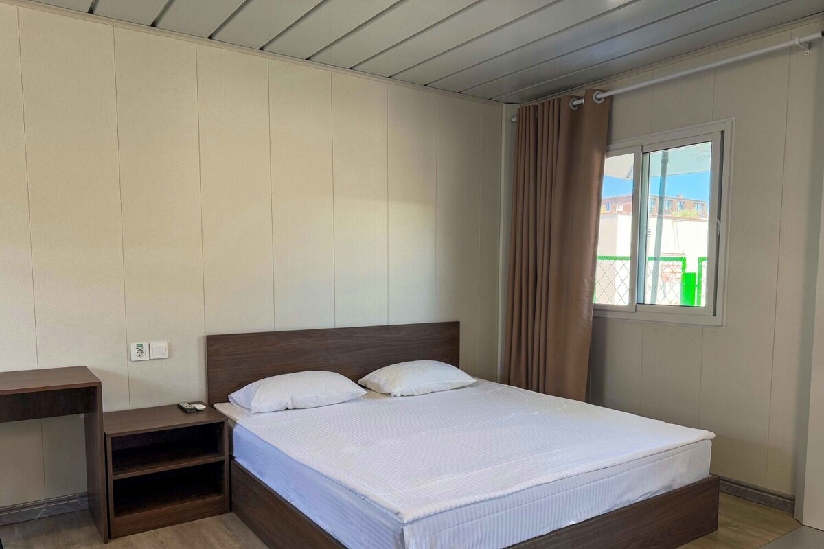 A bedroom with a bed, a bedside table, a window with curtains, and a view of buildings outside.