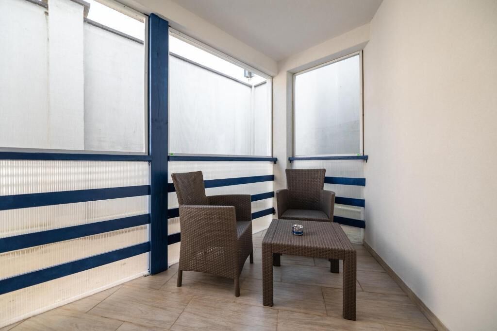 Un balcone con due sedie in vimini, un tavolino, ampie finestre e una ringhiera a righe blu e bianche.