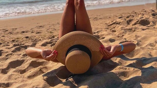 Una persona recostada sobre una playa arenosa, con las piernas levantadas, usando un sombrero de paja y el océano al fondo.