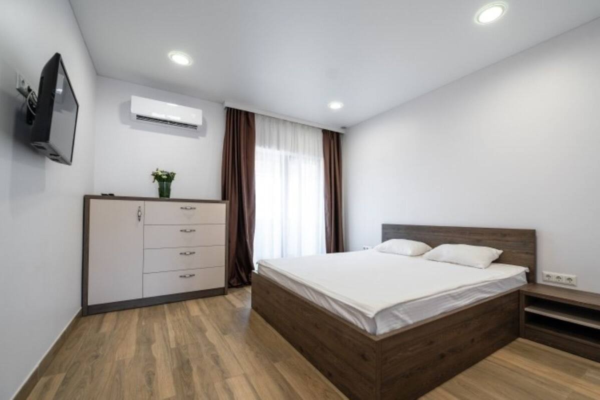 A bedroom with a wooden bed, a dresser, a TV mounted on the wall, and an air conditioning unit.