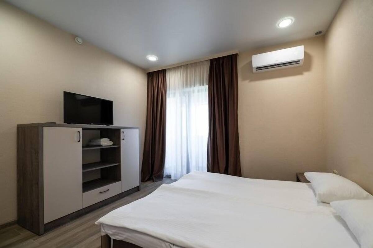 A hotel room with a bed, a TV on a cabinet, an air conditioning unit, and a window with curtains.