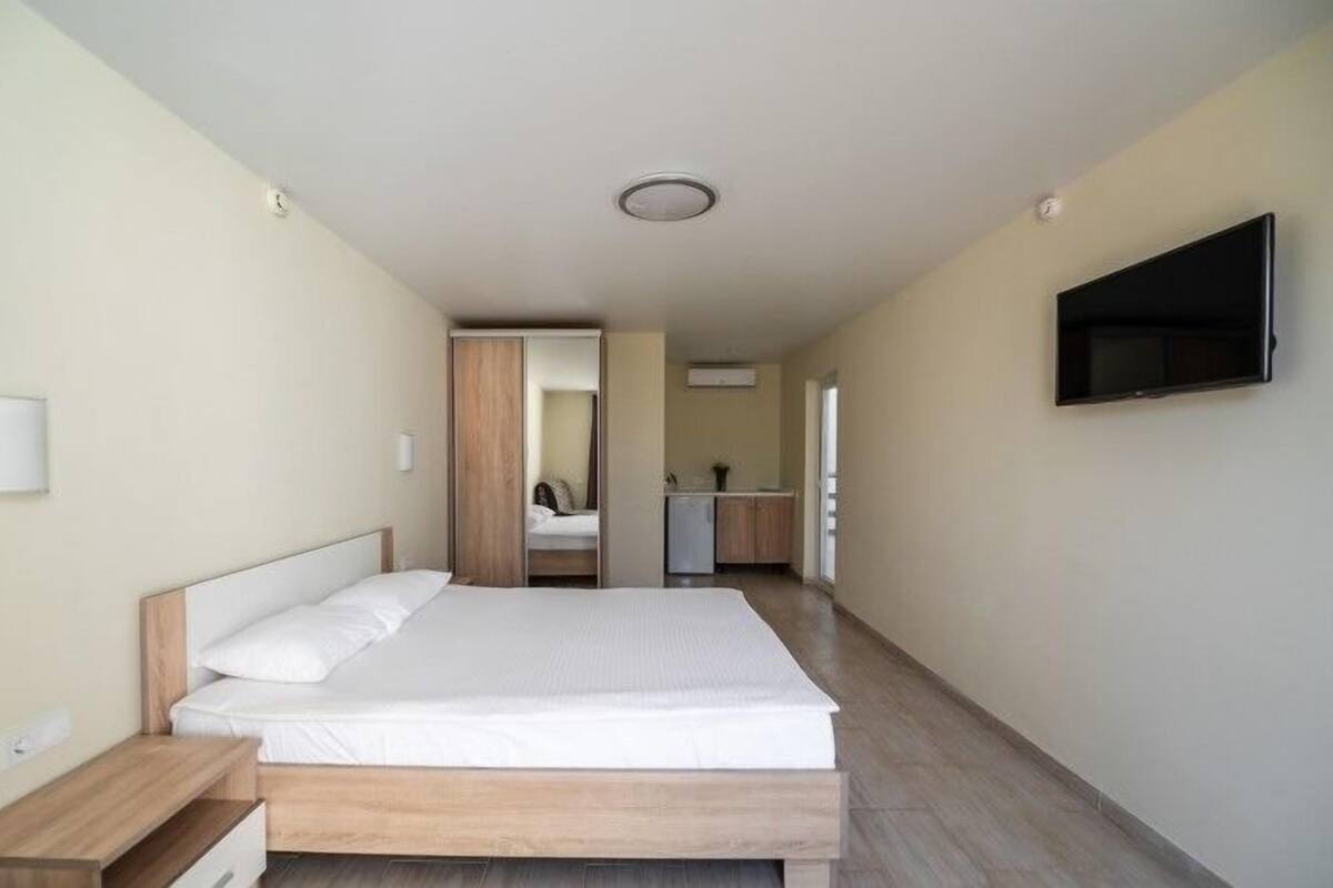 A bedroom with a bed, bedside tables, a TV, and a kitchenette in the background.