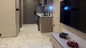 Ett modernt hotellrum med en stor platt-TV, ett litet pentry, ett marmorgolv och en hall som leder till ett annat rum.