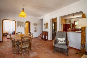 A dining area with a wooden table and chairs, a hanging yellow light fixture, and a wicker chair with a patterned cushion. - Peaceful Cala Vadella Villa with Pool (Sant Josep de sa Talaia)