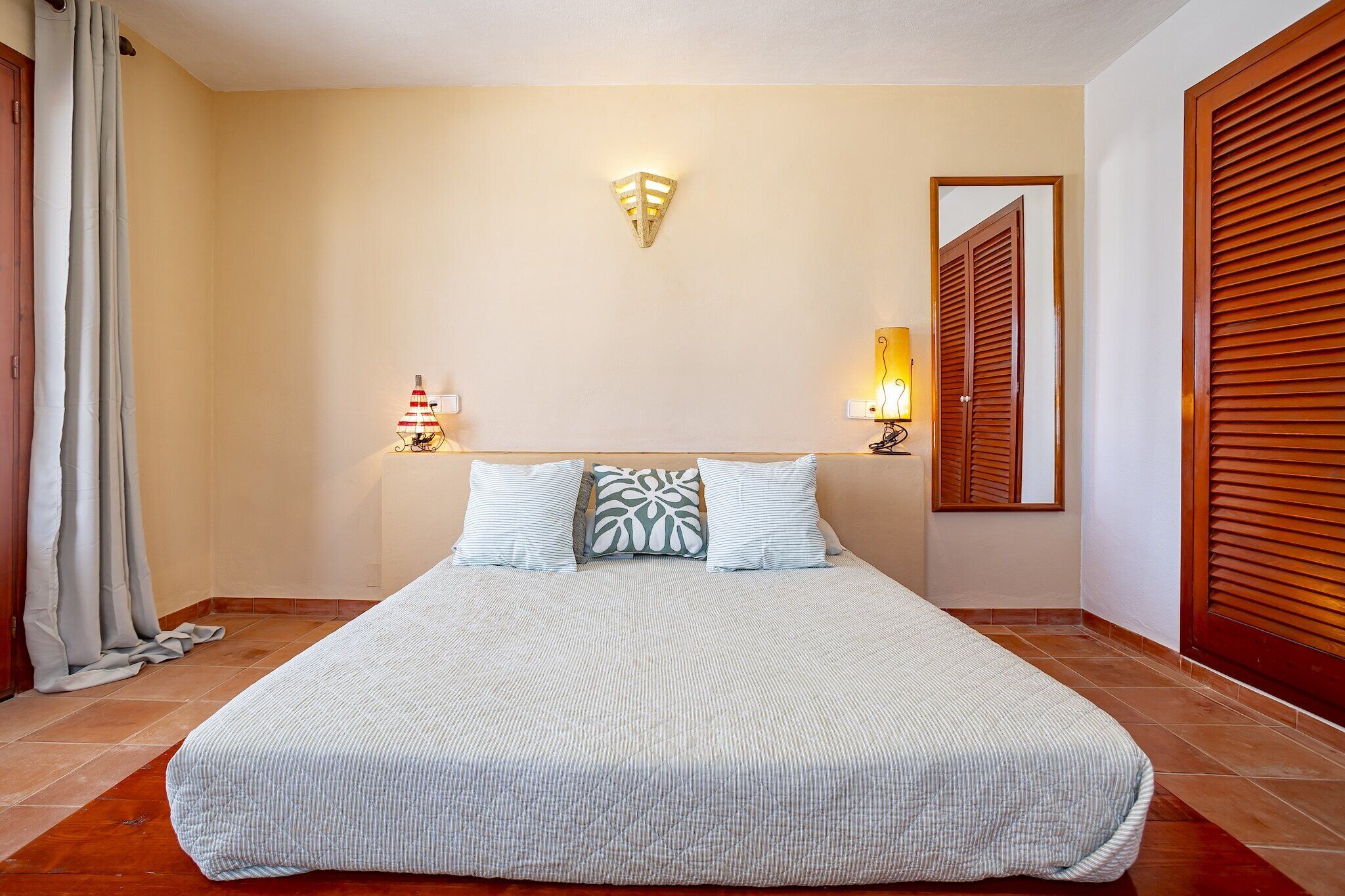 A neatly made bed with a patterned bedspread and pillows, two wall-mounted lamps, and a mirror on the wall.