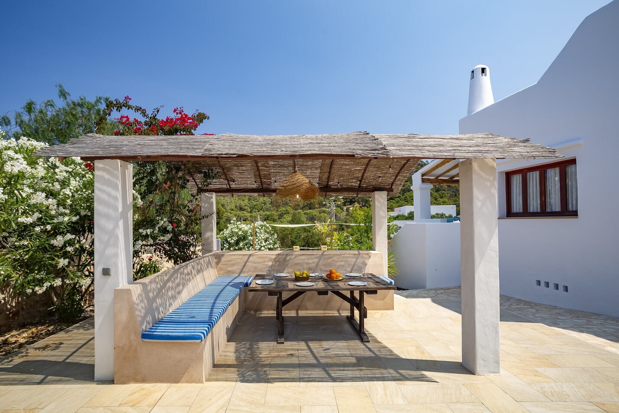 Un espace extérieur aménagé, abrité et doté de tables, prêt pour les repas, entouré d’un mur blanc et d’un jardin planté de fleurs.