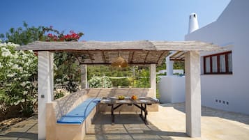 Un espace extérieur aménagé, abrité et doté de tables, prêt pour les repas, entouré d’un mur blanc et d’un jardin planté de fleurs.