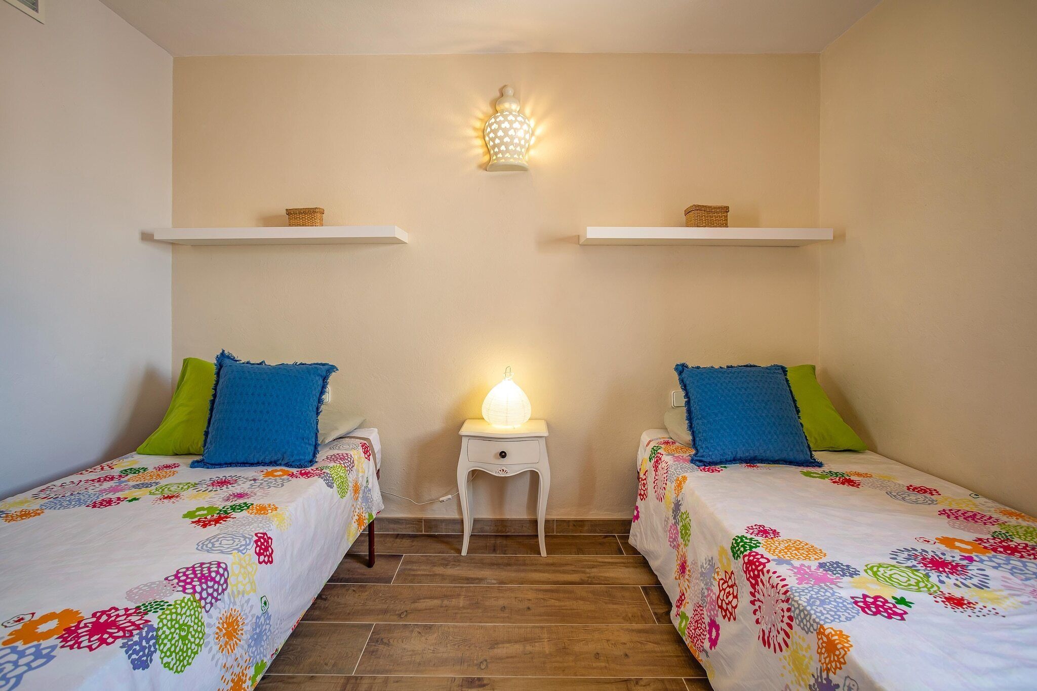 Una habitación con dos camas, cada una con ropa de cama floral y colorida, una mesita de noche pequeña con lámpara y una lámpara de pared.