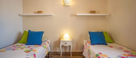 Una habitación con dos camas, cada una con ropa de cama floral y colorida, una mesita de noche pequeña con lámpara y una lámpara de pared.