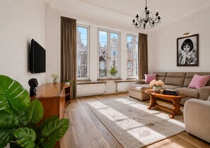 Ein Wohnzimmer mit einer Couch, einem Couchtisch, einem Fernseher und großen Fenstern mit Vorhängen.