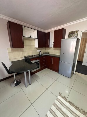 A compact kitchen with dark wood cabinets, a stainless steel refrigerator, and a black countertop. - Cozy, Private & Homey Unit (Randburg)
