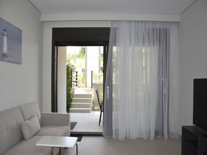 A modern living room with a sofa, a small table, and a TV stand. There is a doorway leading to an outdoor area with steps and greenery. - Two Bedroom Suite Tatiana in Nikiti (Nikiti city)