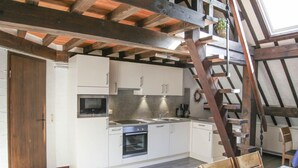 Cocina pequeña con armarios blancos, estufa y lavamanos. Se ve una escalera de madera y una silla.