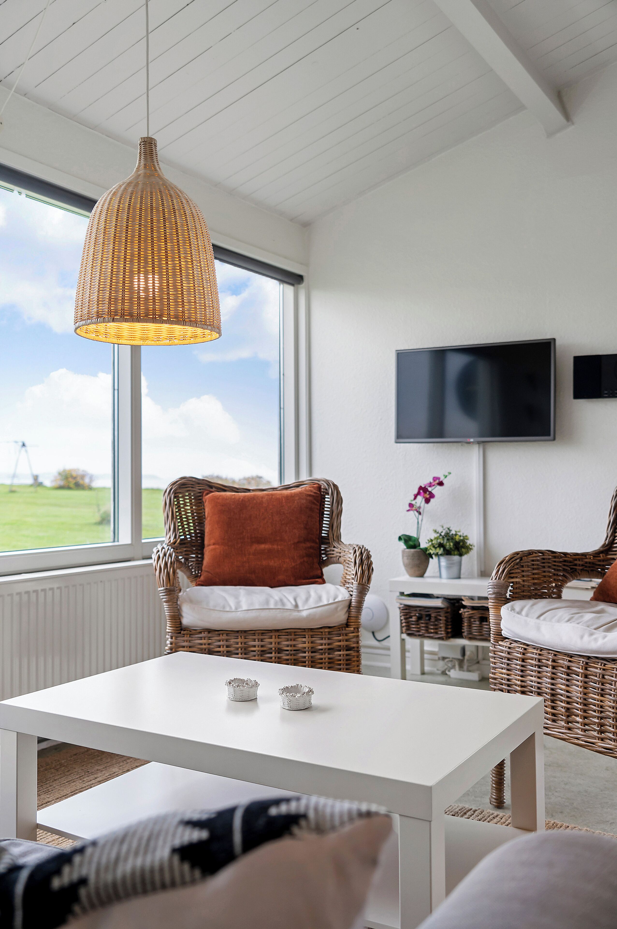 Une pièce meublée de mobilier en osier, avec une table blanche, un téléviseur et une vue sur l’extérieur.