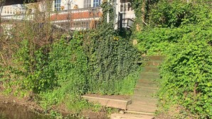 A riverside pathway leading to a building with a red-tiled roof, surrounded by dense greenery.